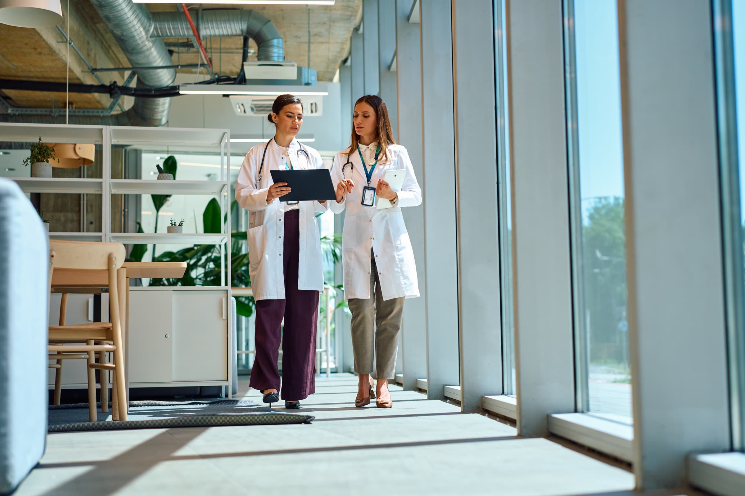 Two clinicians walking a bright clinic hallway while reviewing a tablet